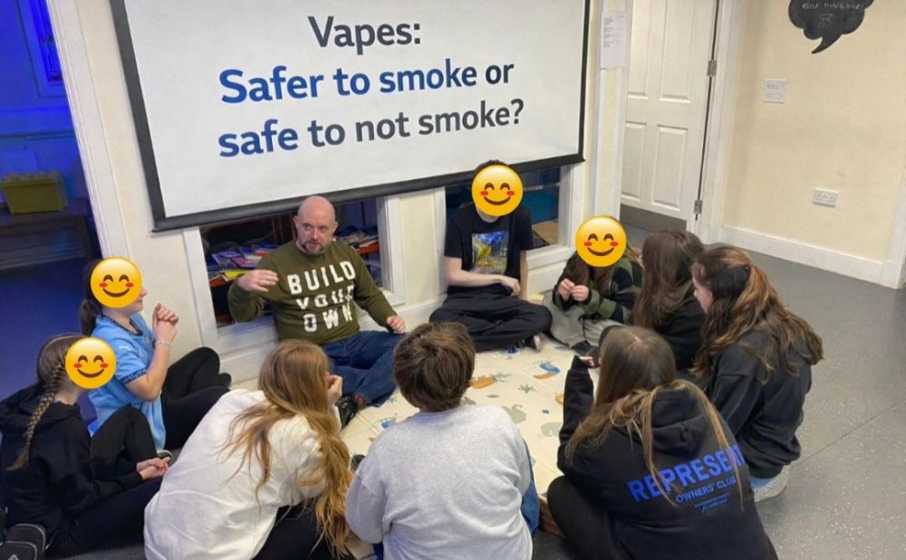 9 young people and one facilitator sitting in a circle engaged in discussion around a smartboard. The smartboard features a question reading: "Vapes: safer to smoke or safe to not smoke?"